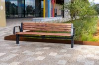 Wooden slat bench with metal frame on a paved plaza, with a glass-front building and colorful vertical poles in the background.