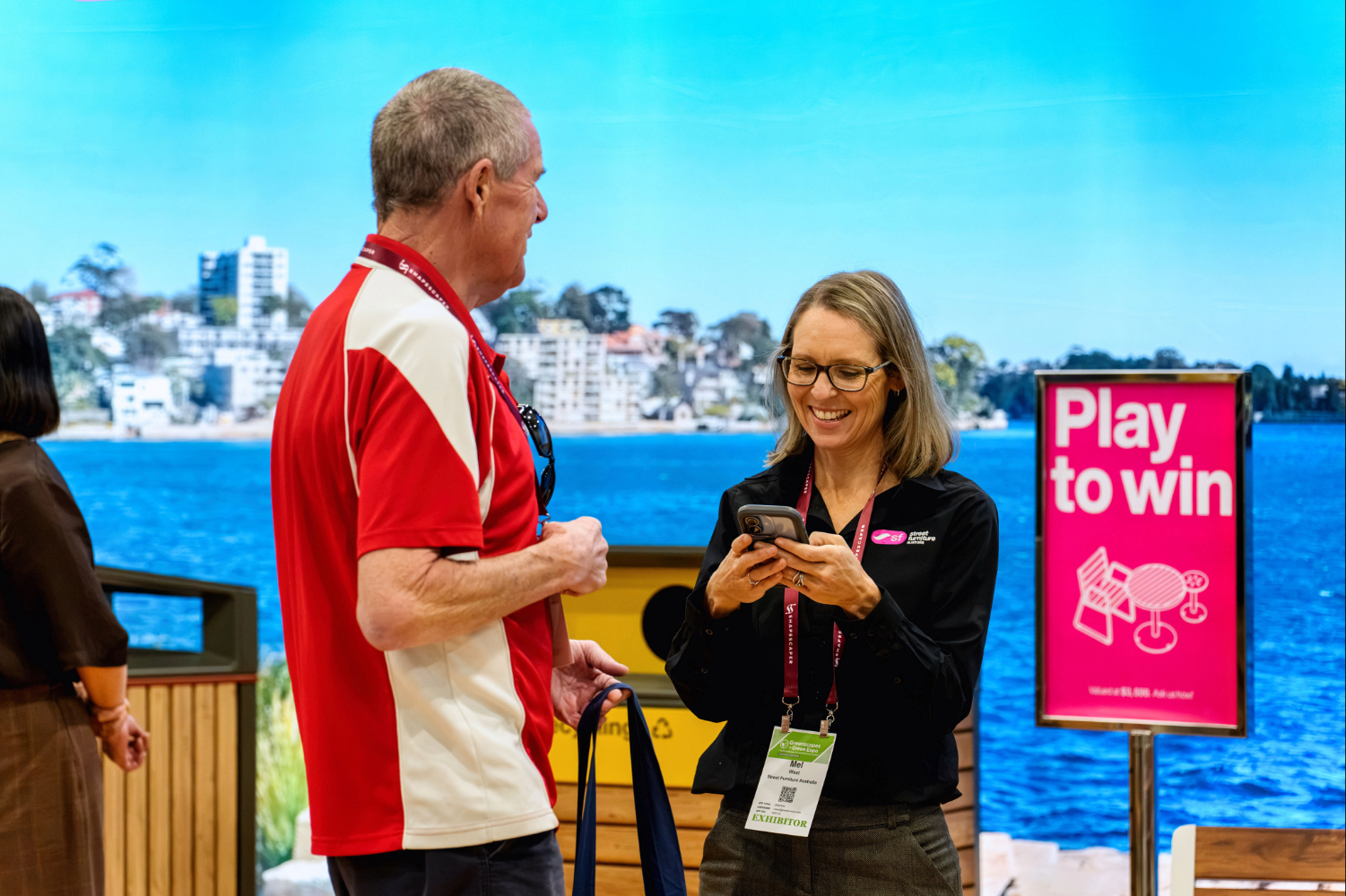 Two adults chat at an expo booth by a seaside backdrop; the woman shows her phone while the man in a red-and-white polo listens.