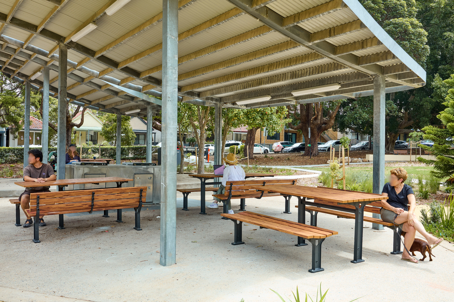 Johnson Park. People sit at wooden picnic tables under a metal-roofed pavilion in a park, with trees and parked cars in the background.