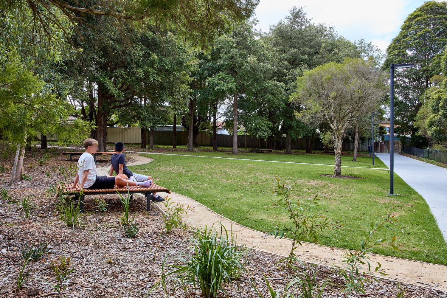 Johnson Park, Two boys sit on a wooden slatted bench beside a curved mulch bed in a park, with tall trees and a green lawn in the background.