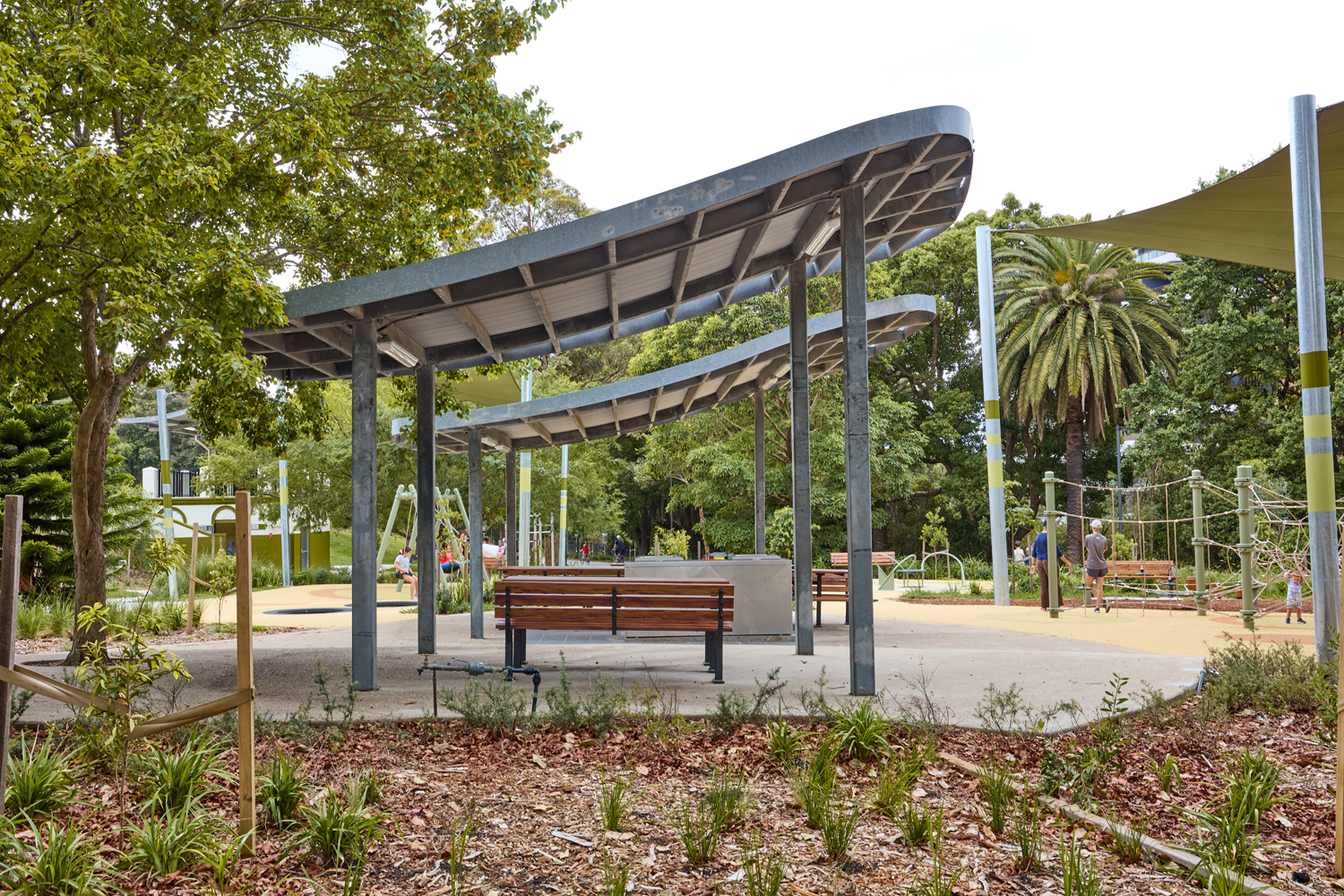 Johnson Park. Curved metal shade structures over a seating area in a sunlit park, with benches and kids playing in the distance.