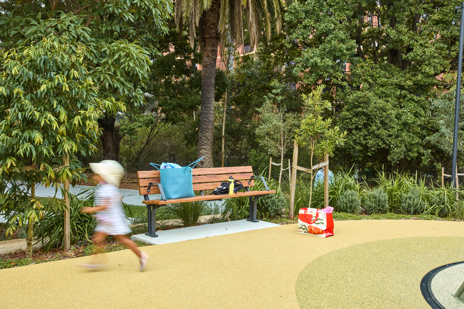 Johnson Park. A wooden park bench under trees with a blue tote bag and a water bottle, while a child runs past in a blurred motion.