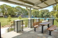 Johnson Park. Covered outdoor seating area with wooden picnic table and benches overlooking a grassy park and basketball court in the distance.