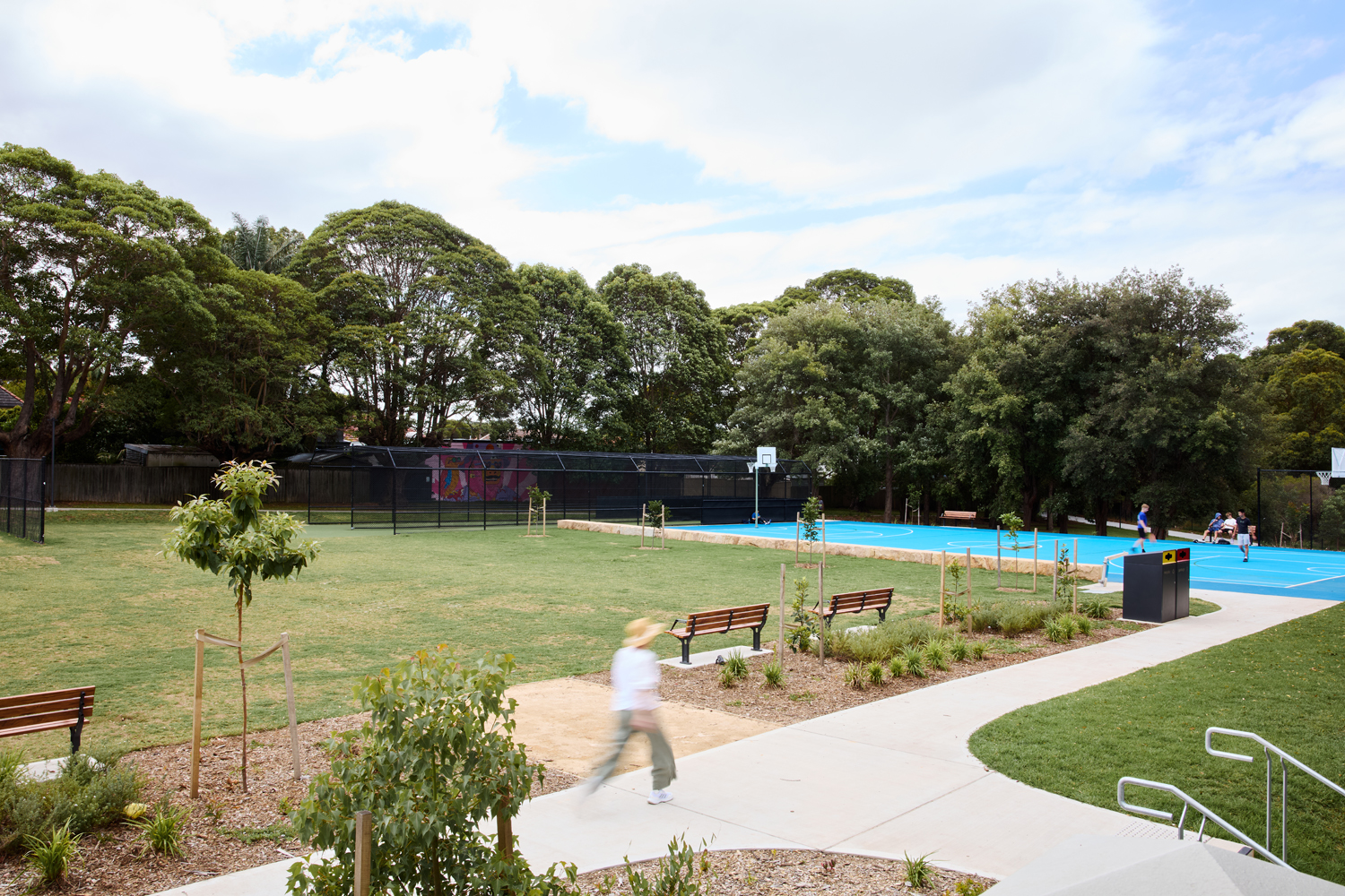 Johnson Park. Park scene with a bright blue basketball court, surrounding trees, and benches along a paved path; a person is walking in the foreground.