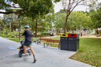 Johnson Park. Blurred cyclist and stroller pass by a park bench near a two-bin recycling/garbage station, with a playground and people in the background.