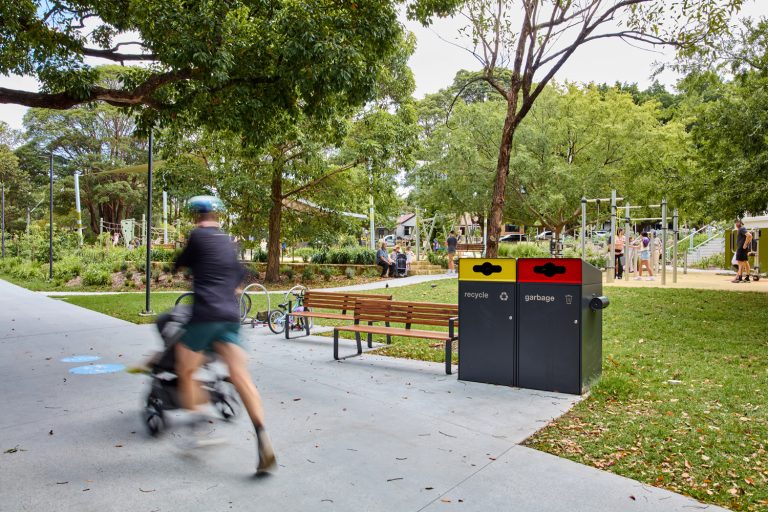 Johnson Park. Blurred cyclist and stroller pass by a park bench near a two-bin recycling/garbage station, with a playground and people in the background.