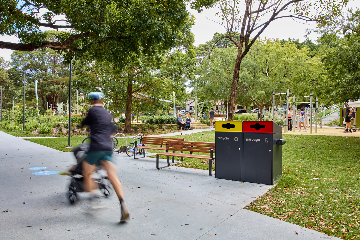 Johnson Park. Blurred cyclist and stroller pass by a park bench near a two-bin recycling/garbage station, with a playground and people in the background.