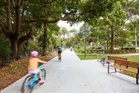 Johnson Park. People riding bikes on a sunny park path lined with trees and a bench to the right