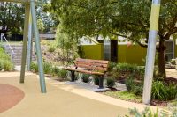 Johnson Park. Empty wooden bench with metal supports on a concrete pad in a sunlit park, surrounded by greenery and a tree overhead
