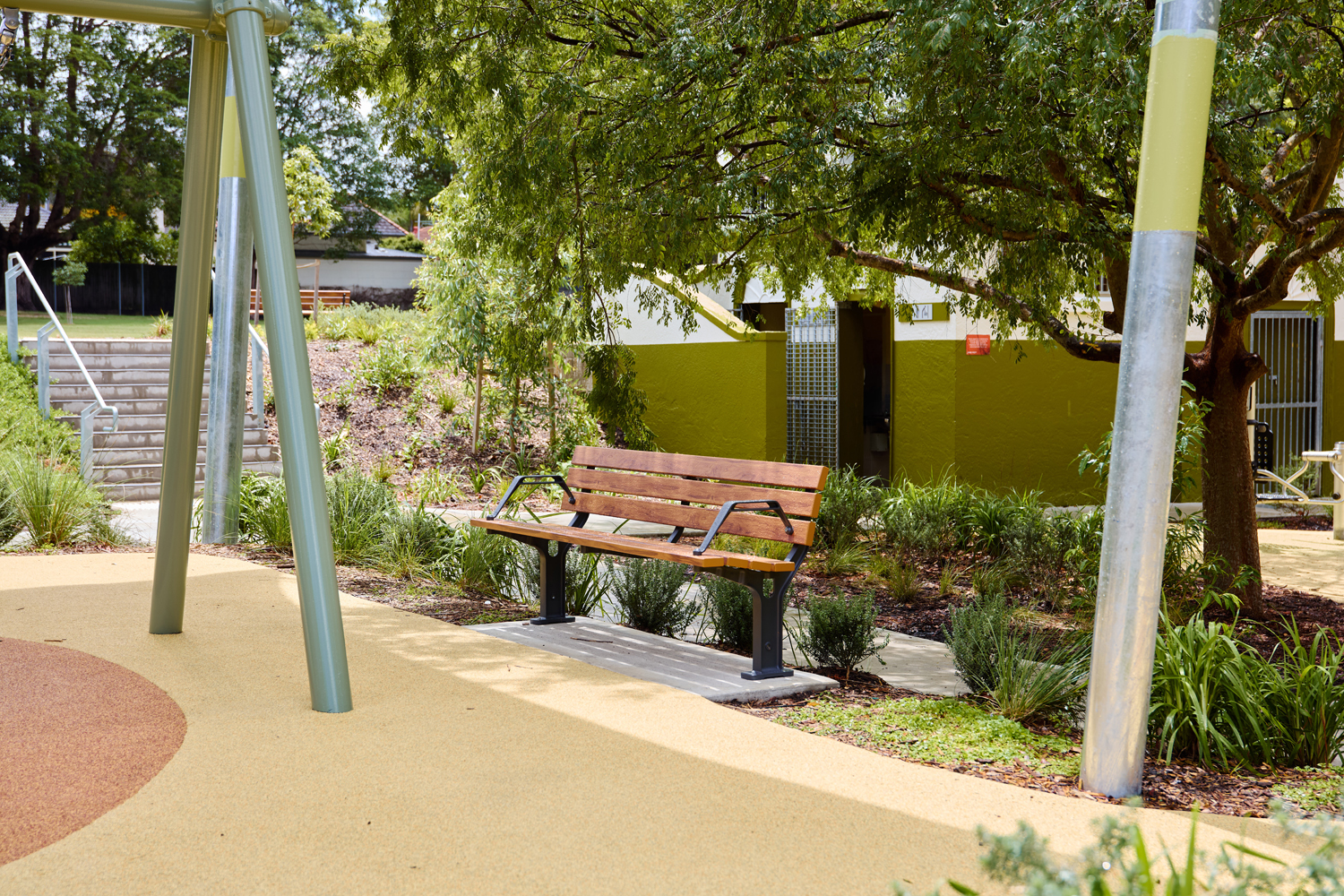 Johnson Park. Empty wooden bench with metal supports on a concrete pad in a sunlit park, surrounded by greenery and a tree overhead