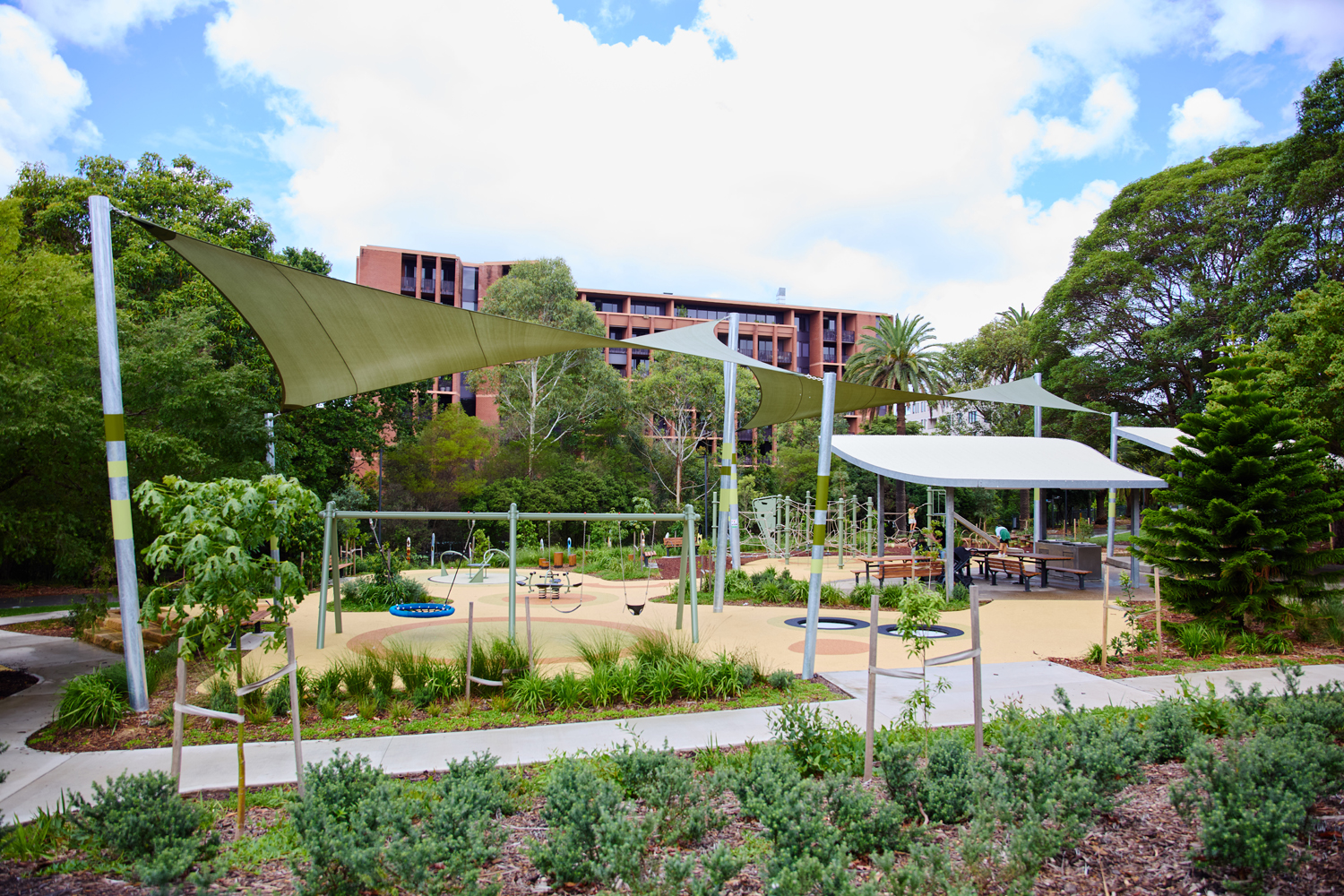 Johnson Park. Children's playground with climbing structures and rubberized surface under green shade sails in a sunny park area.