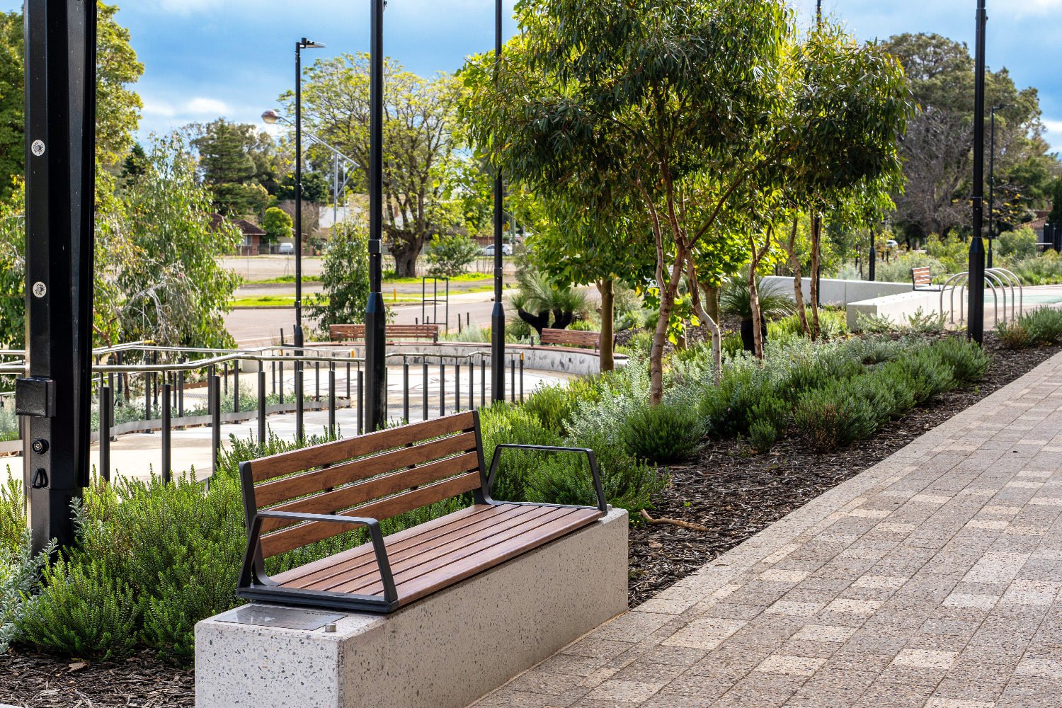 A wooden slat bench with metal arms on a concrete base beside a paved walkway in a park setting with trees and shrubs nearby.