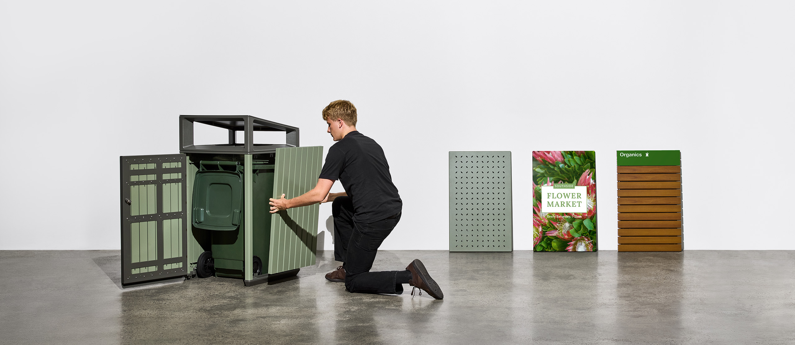 Man kneels to position green corrugated panels beside a large green waste bin in a bright studio setting.