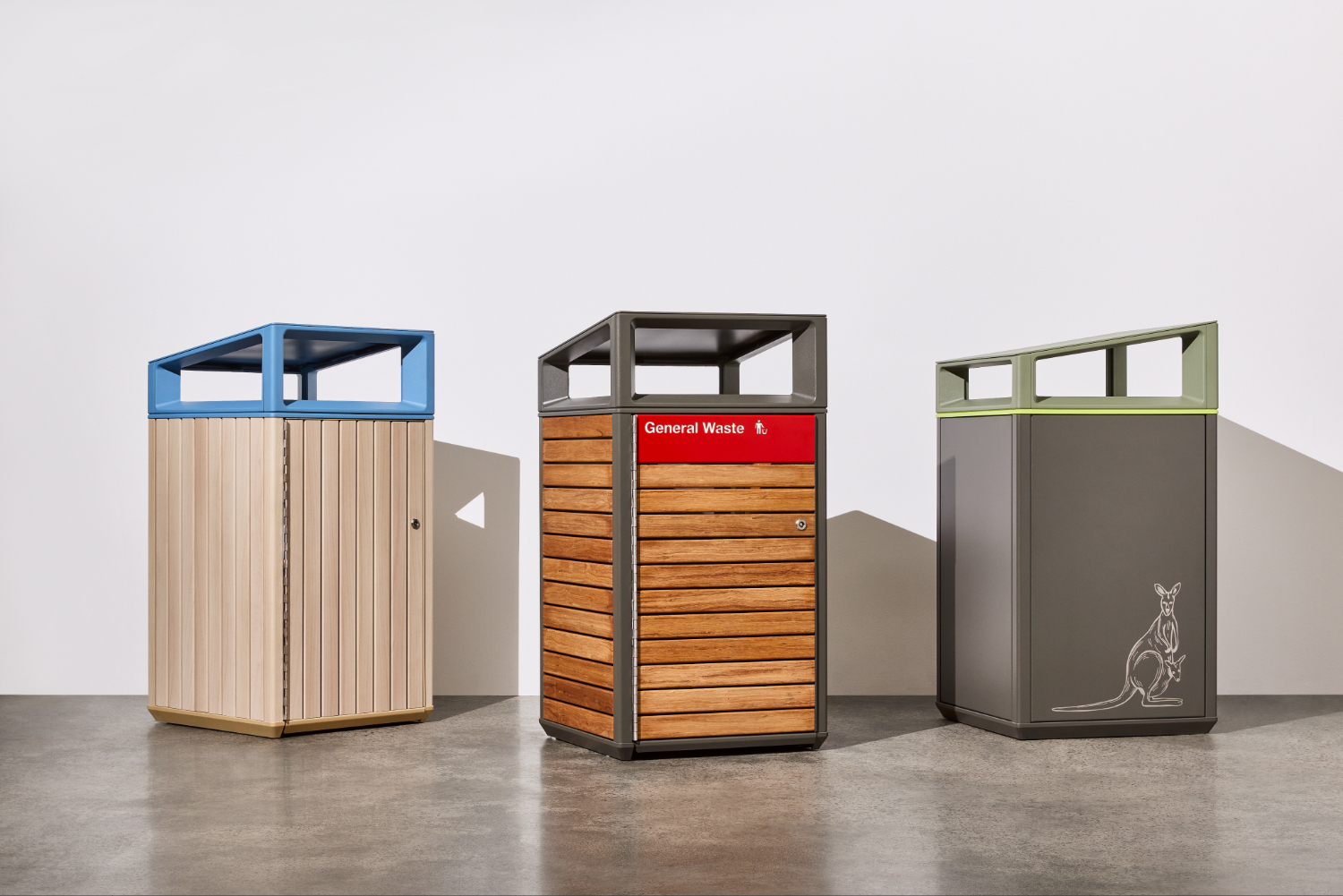 Three outdoor waste bins in a row: left a light wood-slatted bin with a blue frame, center a wood-and-gray bin labeled 'General Waste', right a dark gray bin with a kangaroo illustration.