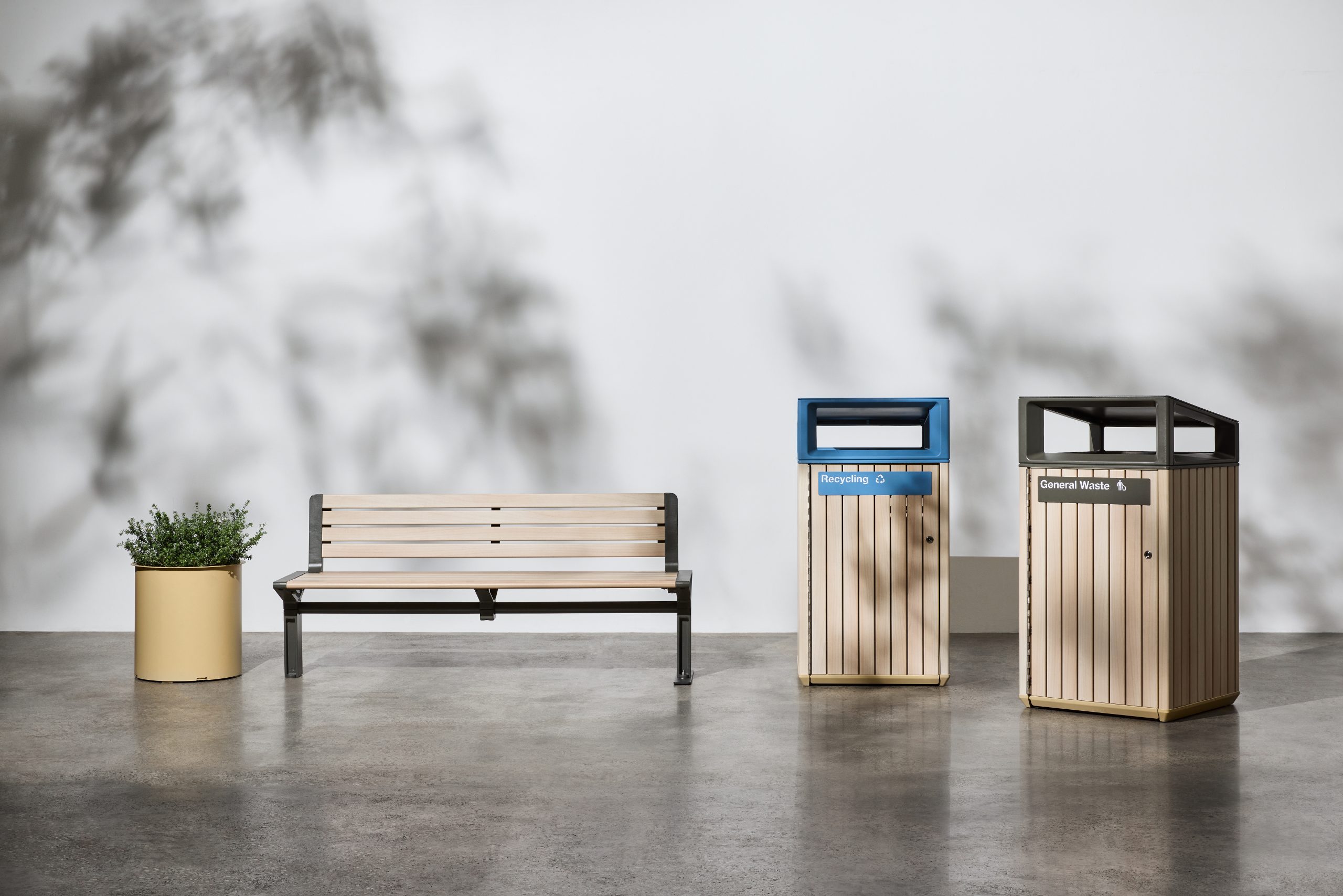 Wooden bench beside two labeled waste bins (Recycling and General Waste) in a clean indoor setting, with a potted plant to the left.