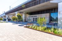 Armadale Station entrance under an elevated rail line, with benches, trees, and a paved plaza. Readable station signage nearby.