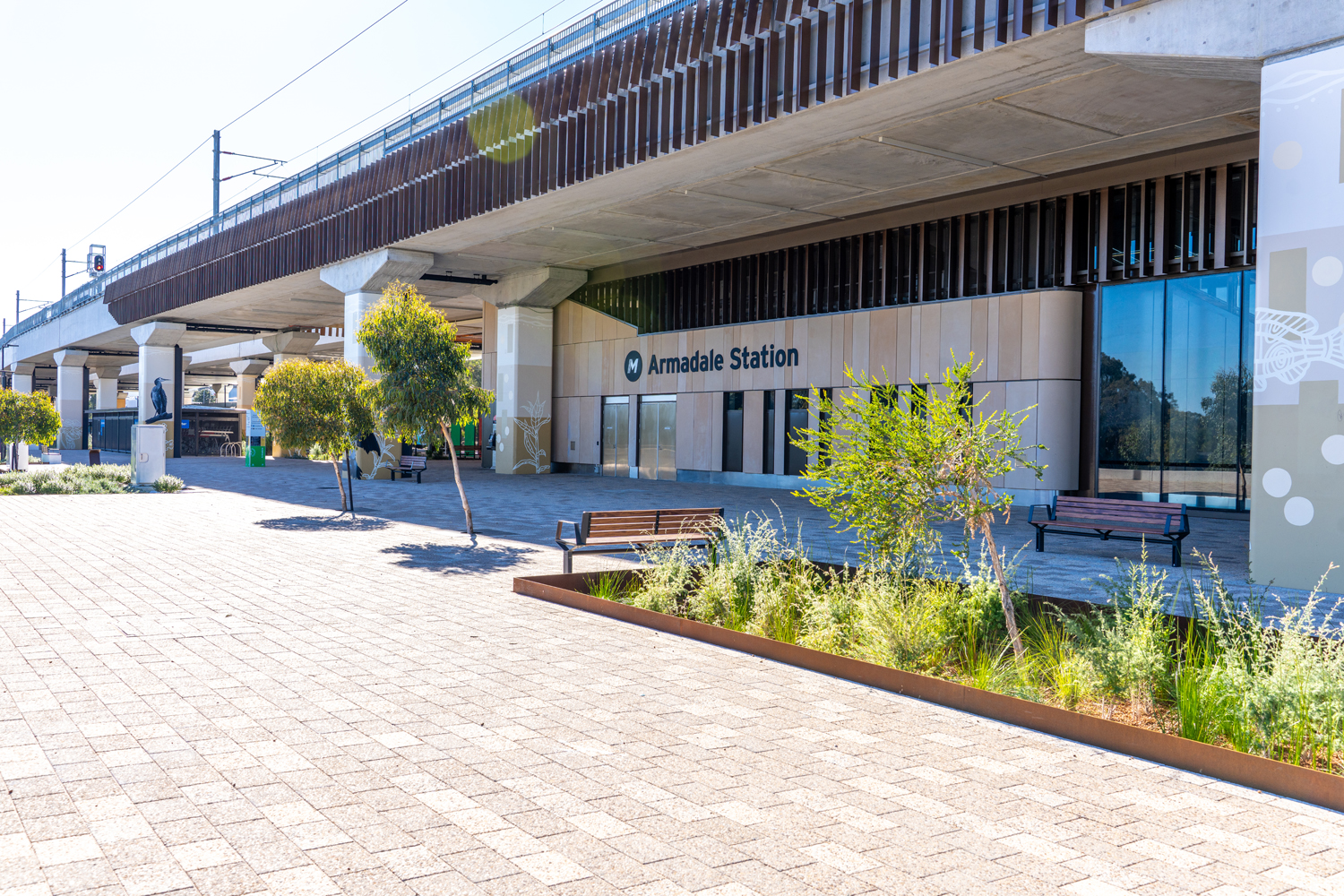 Armadale Station entrance under an elevated rail line, with benches, trees, and a paved plaza. Readable station signage nearby.