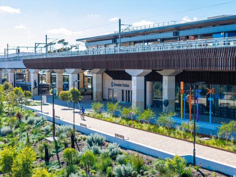 Armadale Station plaza with elevated rail, colorful art poles, and a cyclist on the pedestrian path amid landscaping.
