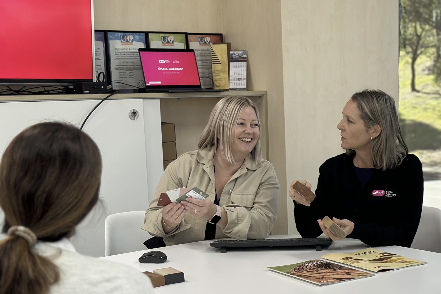 Two women sit at a white table comparing color swatches while a third person watches, smiling during a product consultation in a showroom setting.