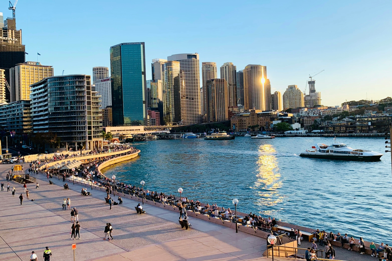 Bustling waterfront promenade with a blue harbor, high-rise skyline, and people strolling along the edge.
