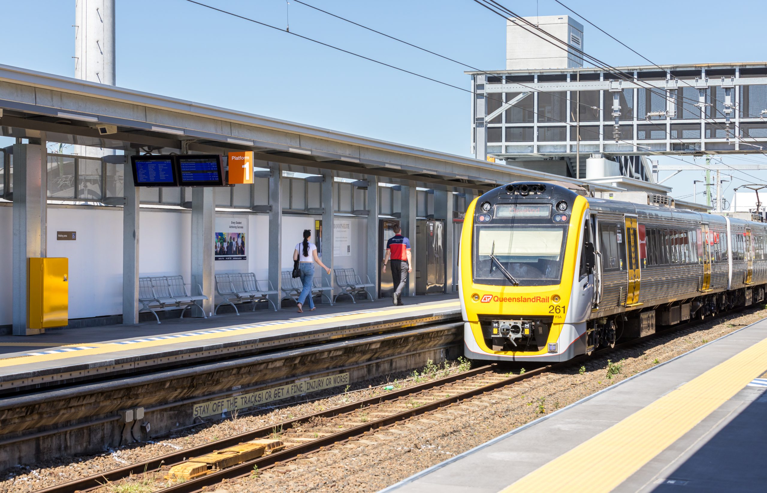 Yellow diesel train at a platform with passengers waiting under a canopy and digital timetable boards in view.