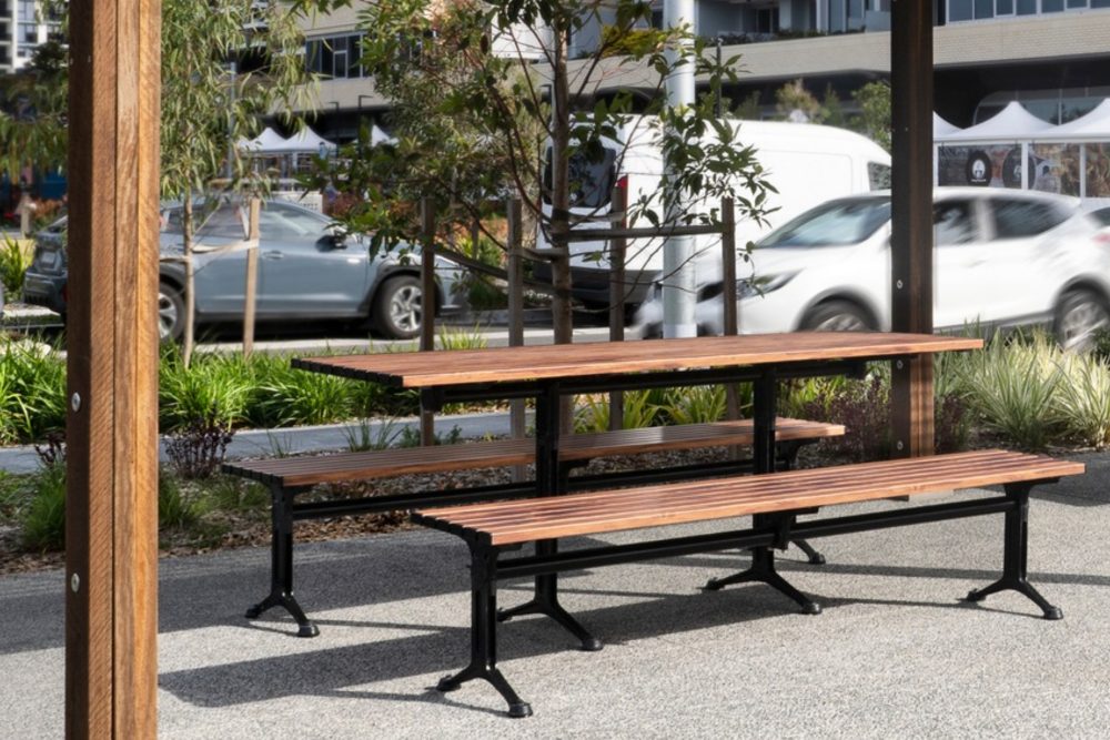 Untitled Design (16) - Street Furniture Australia Wooden outdoor benches on metal frames in a landscaped urban park with trees and event tents in the background.