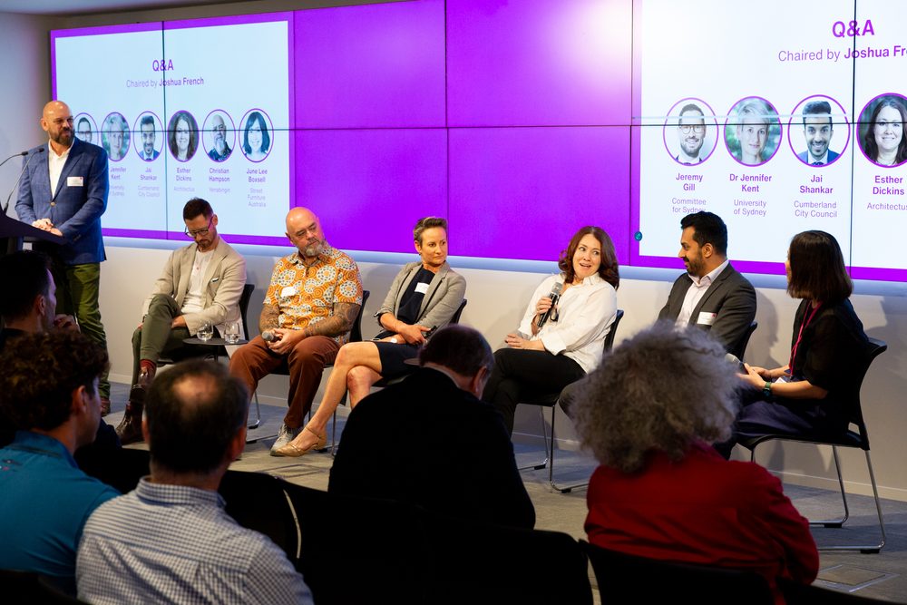 Panel discussion on stage with six seated speakers during a Q&A, audience in foreground; large purple screens behind show speaker profiles and titles.