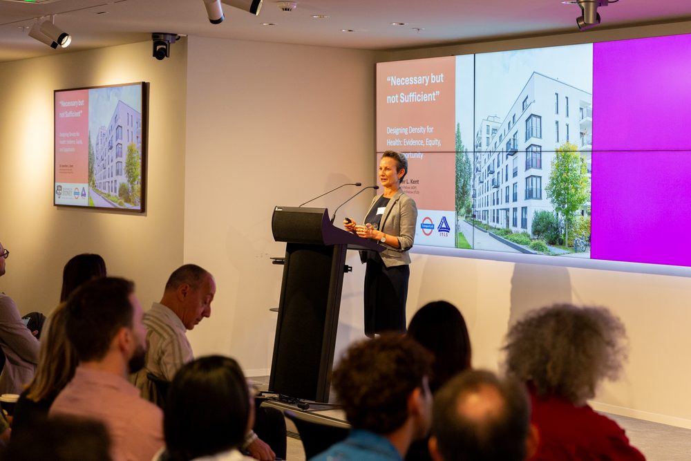 Professional woman presenting at a black podium in a conference room; a large multi-panel screen behind her displays a slide titled 'Necessary but not Sufficient' with an image of a modern apartment building and a purple panel to the right; audience seated and listening.