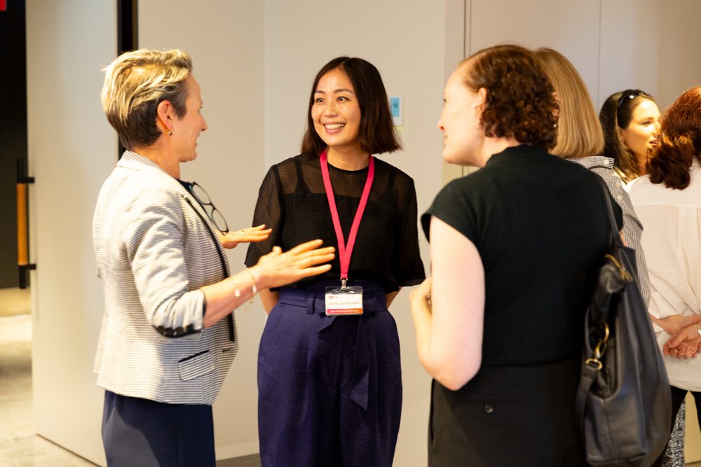 Smiling group of professionals chatting in a hallway; woman in center wears pink lanyard and name badge.
