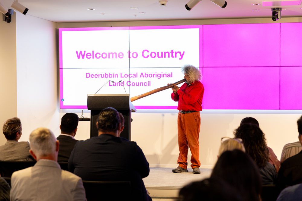 Man with gray curly hair plays a didgeridoo on stage, wearing a red shirt, in front of a pink display and seated audience attendees. (Welcome to Country scene)