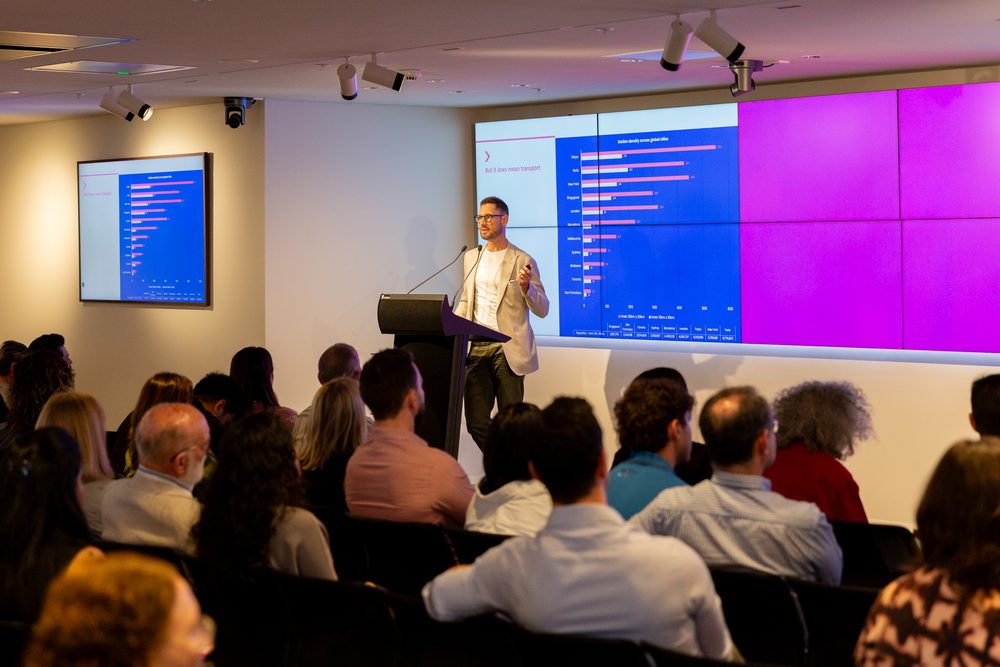 Male presenter in a light blazer at a lectern, presenting with blue charts shown on a large screen behind him.