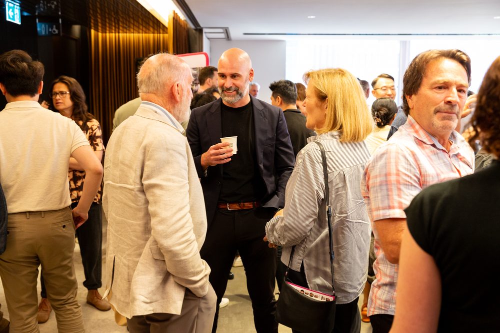 Professionals mingle at an indoor networking event, conversing in small groups near a wooden wall.