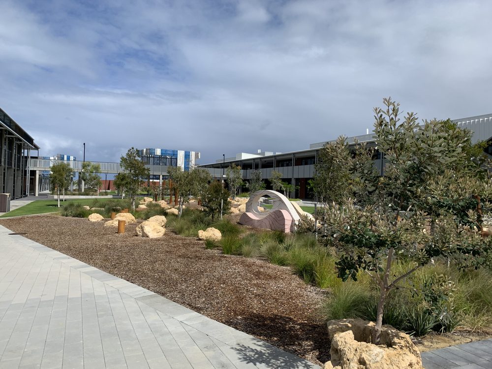 Campus courtyard with a paved walkway, modern buildings, and green landscaping under a cloudy blue sky.