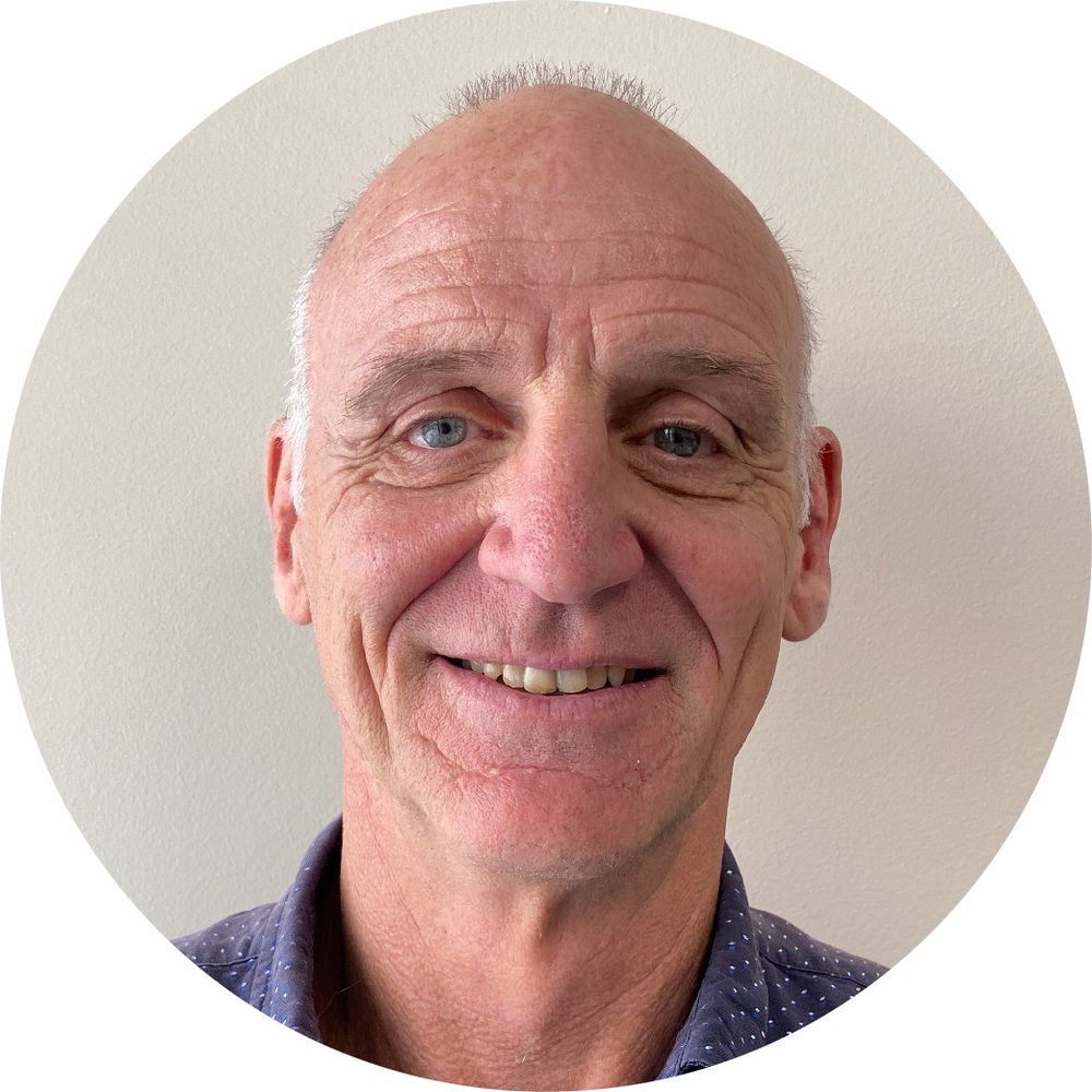Smiling older man with blue eyes and balding gray hair, wearing a navy polka-dot shirt against a light wall.