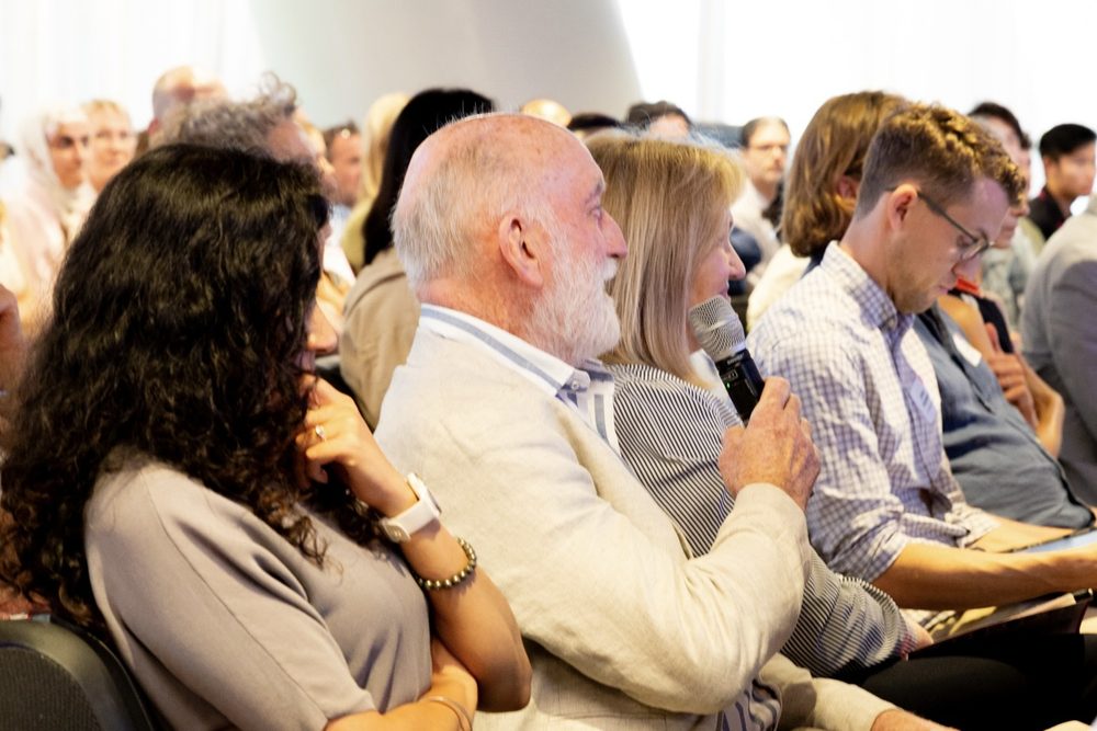 Audience at a conference or seminar; an older man with a white beard holds a microphone while seated among other listeners.