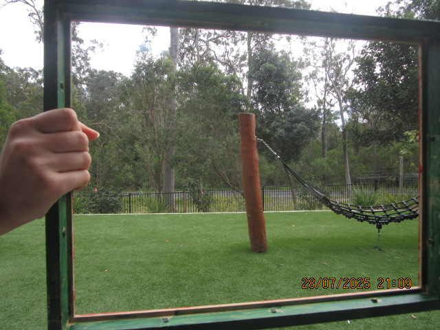Hand holds a green frame that frames a playground with a hammock tied to a wooden post on artificial grass